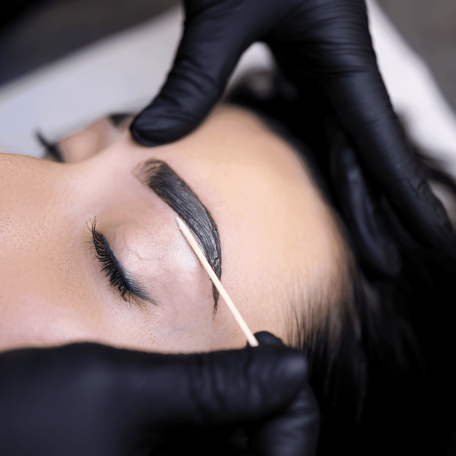 Eyebrow shaping in progress with a wooden stick, performed by a person wearing black gloves.