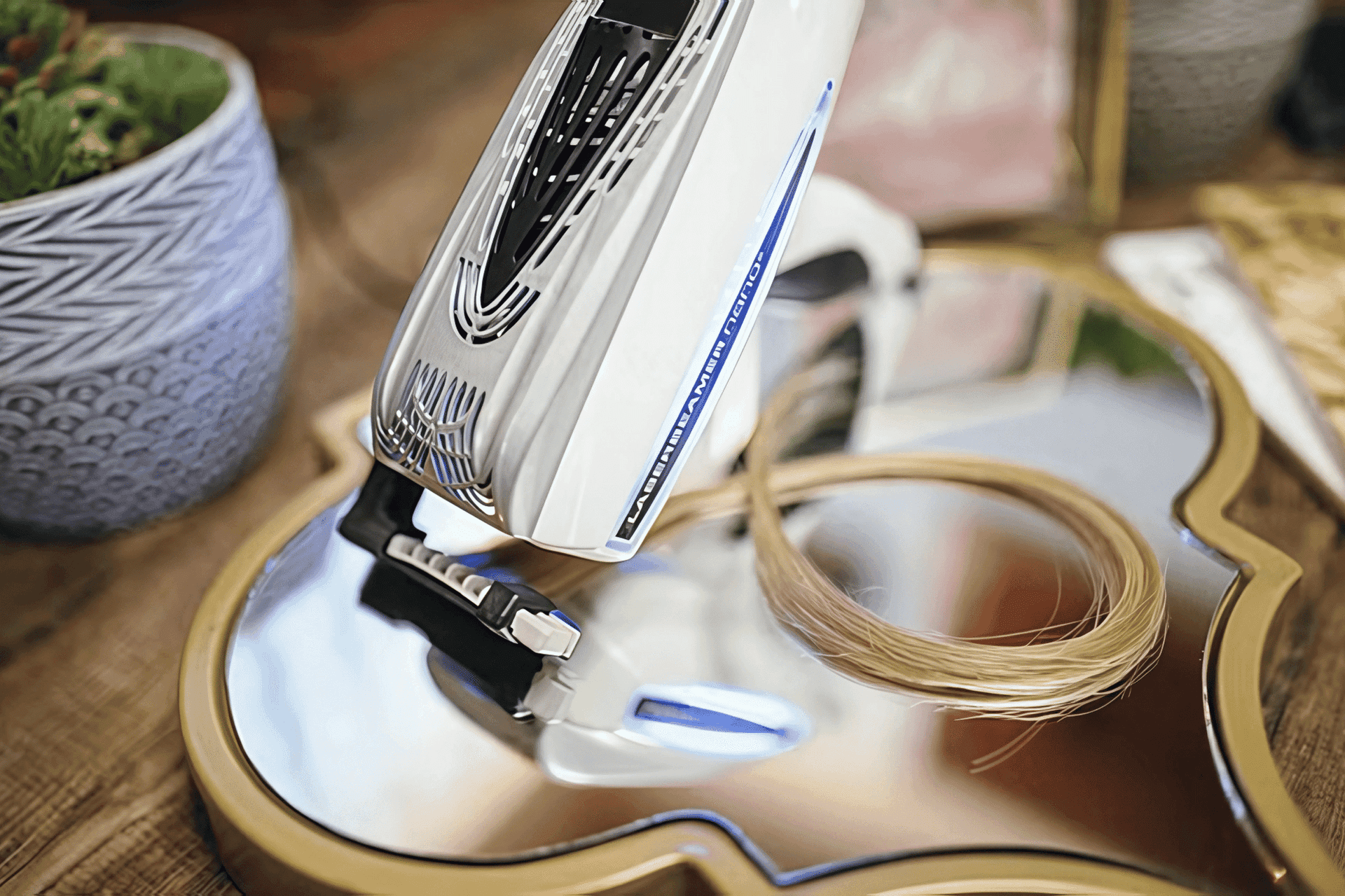 Haircare device lies on a reflective surface with a lock of hair, plants, and a comb nearby.